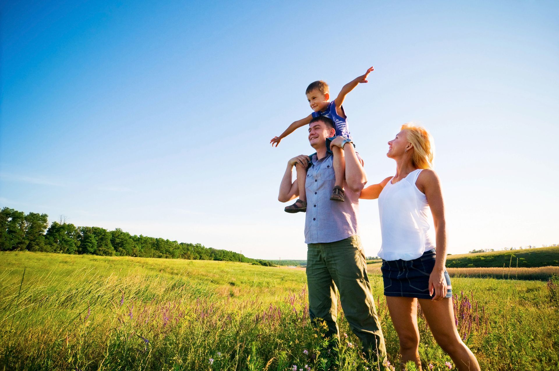 A family standing in a grassy field on a sunny day, with a child sitting on a parent's shoulders with arms outstretched.