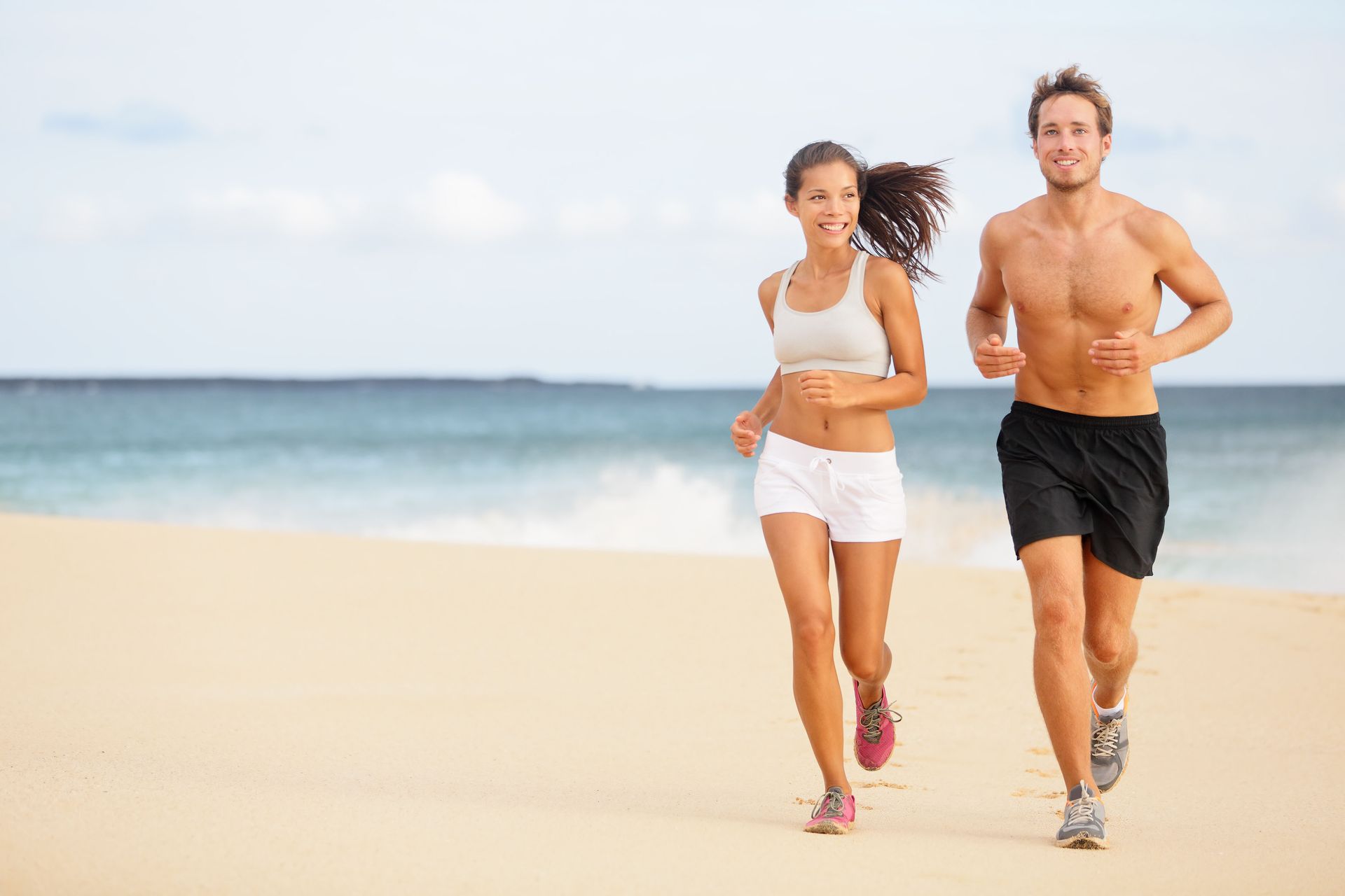 A fit couple runs along a sandy beach with the ocean in the background.