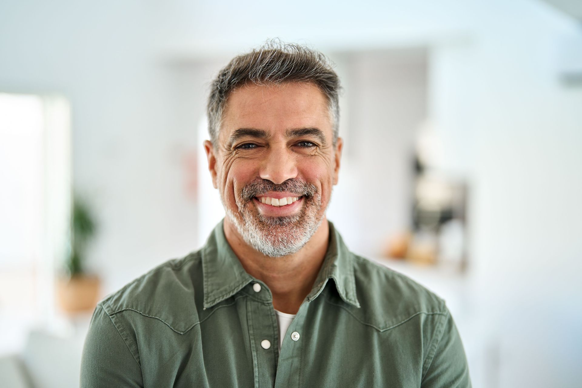 A smiling person with a short beard wears a green button-down shirt, captured in a softly blurred home setting.
