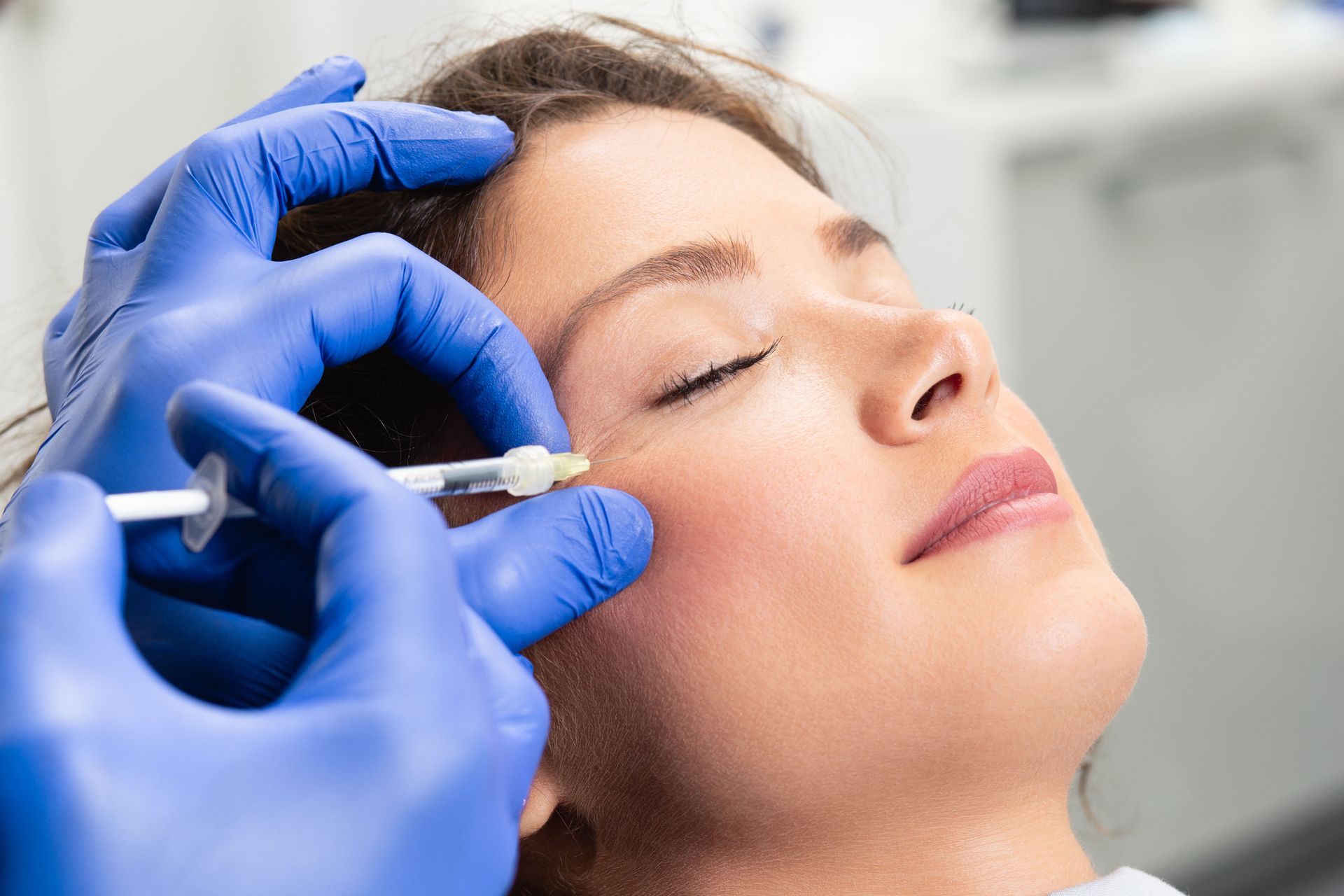 A medical professional in blue gloves administers a facial injection to a person resting with their eyes closed.