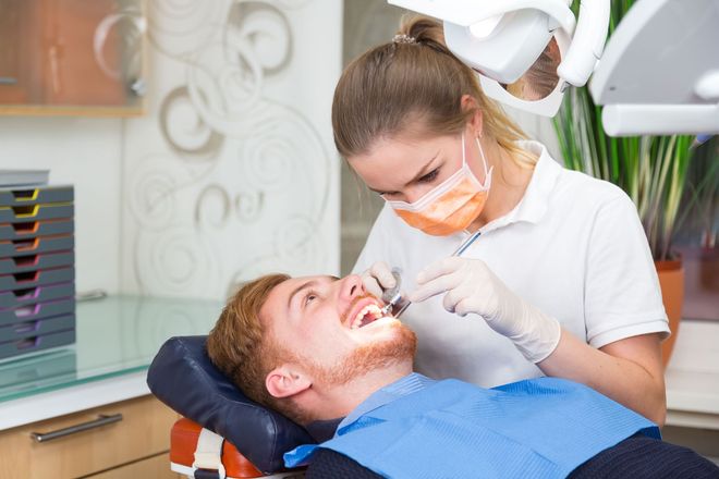 Dentist treating a patient in a dental chair, using tools while wearing a mask and gloves.