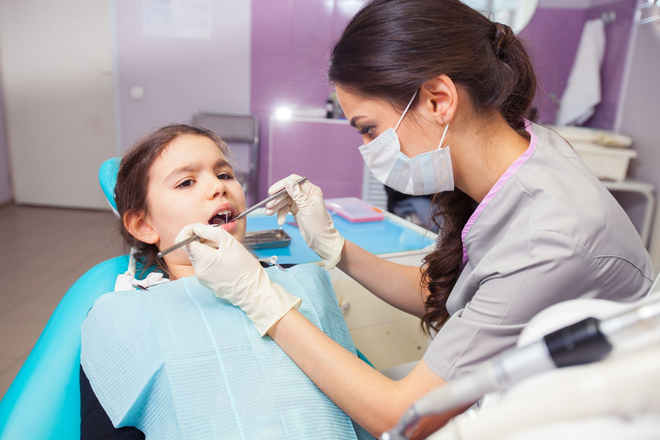 Dentist examining a child patient in a dental chair, wearing gloves and mask.