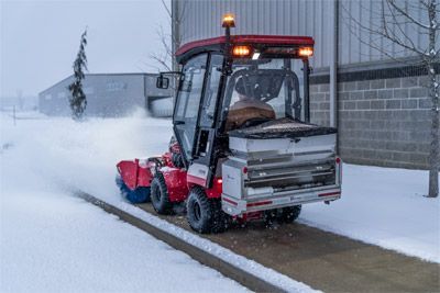 Red snowblower clearing snow from a sidewalk. Salt spreader on the back. Winter setting.