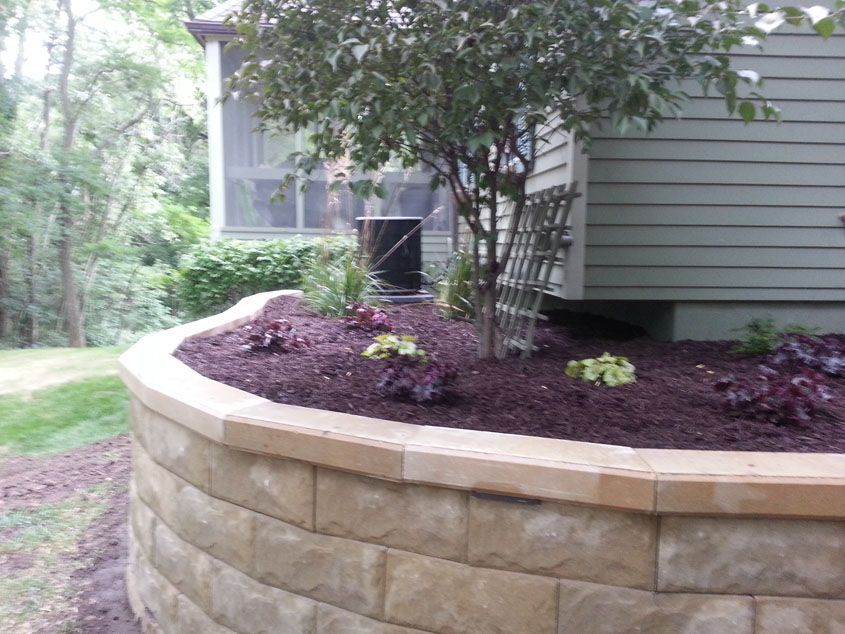 Tan stone retaining wall with a landscaped bed of dark mulch, plants, and small tree next to a house