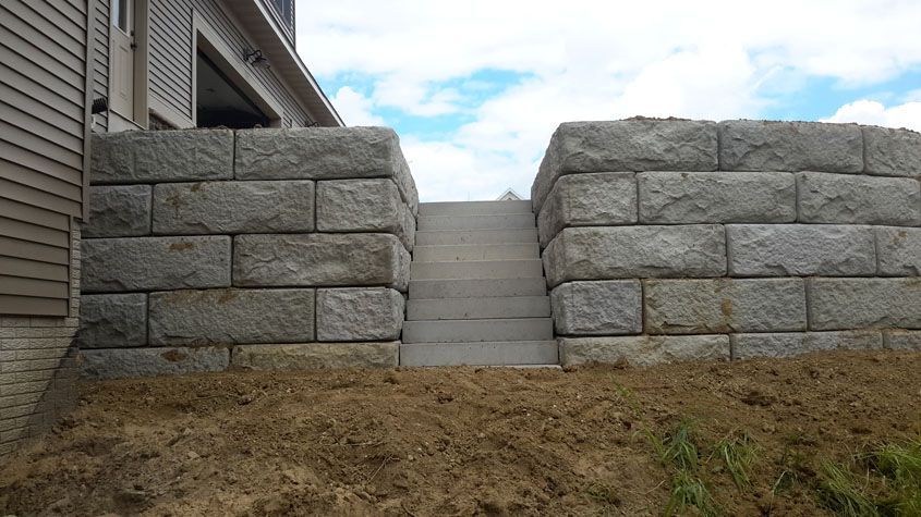 Stone block walls flank concrete stairs leading up to a building, set on a grassy hill, with blue sky