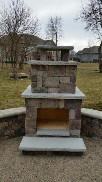 Outdoor brick fireplace with a small chimney, set on a concrete patio in a backyard