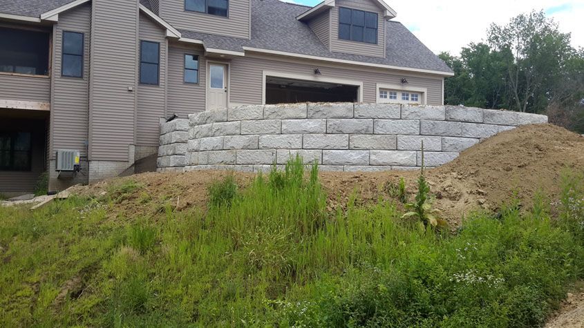 Stone retaining wall in front of a beige house with a garage, overgrown grass in foreground