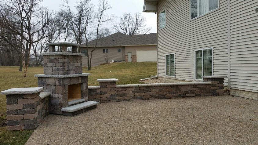 Outdoor patio with a stone fireplace and retaining wall beside a house