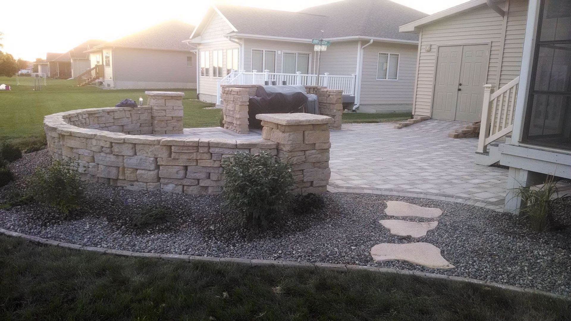 Stone patio with built-in grill and curved seating area in a backyard, surrounded by landscaping