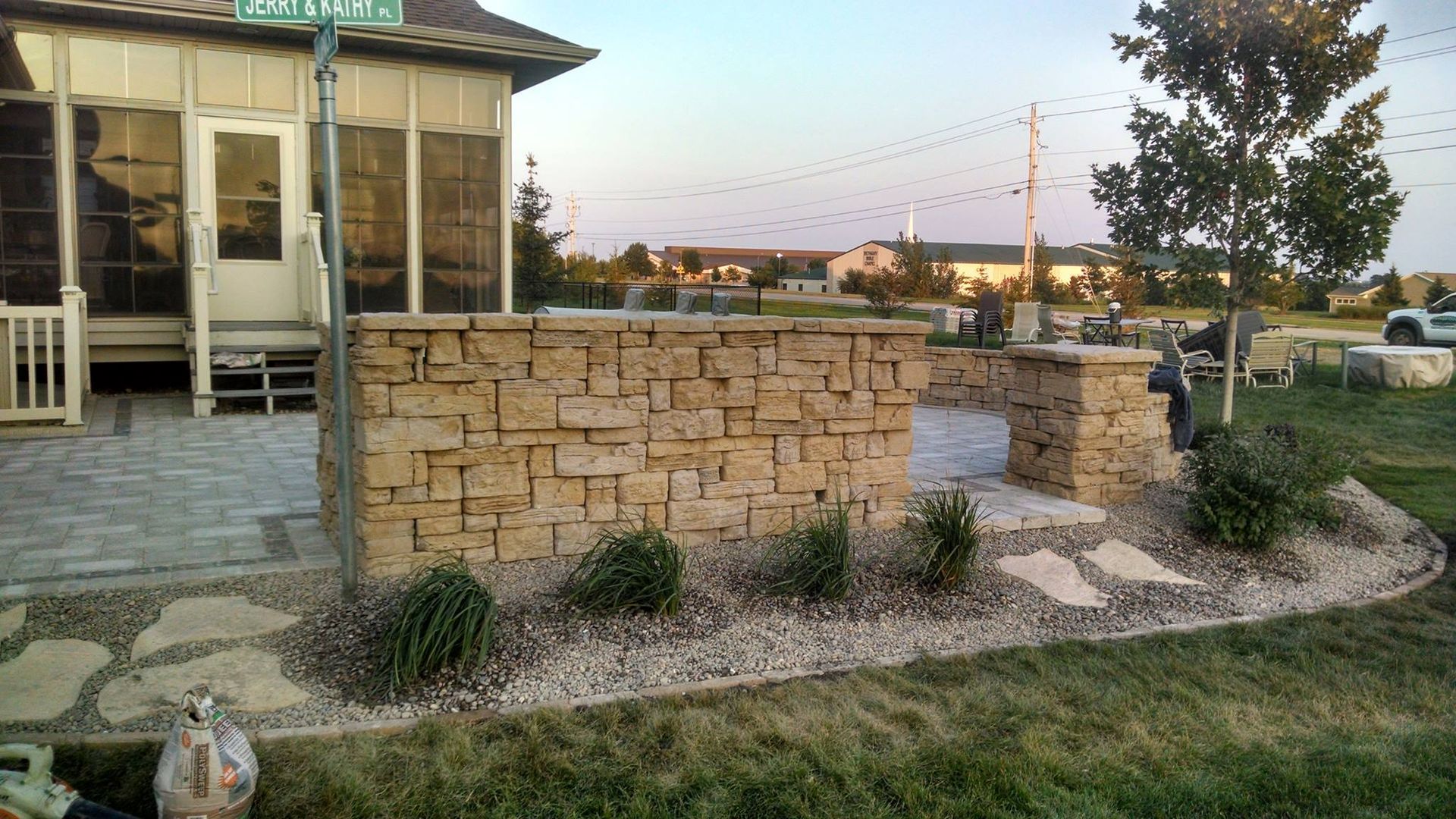 Stone wall with archway on a patio, landscaping with greenery, in front of a house with porch