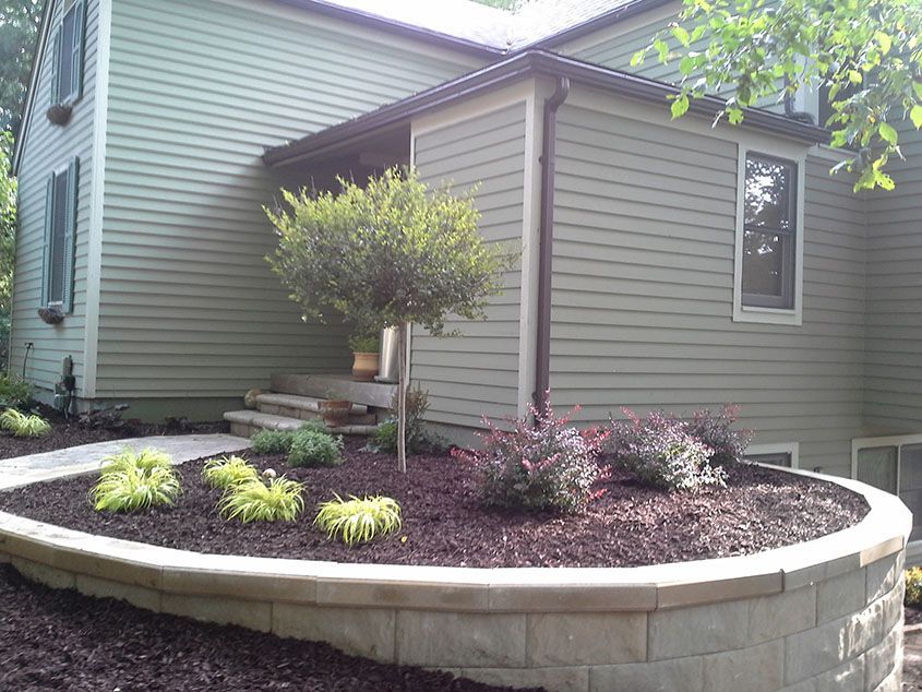 House with a raised garden bed filled with plants, a small tree, and stone retaining wall