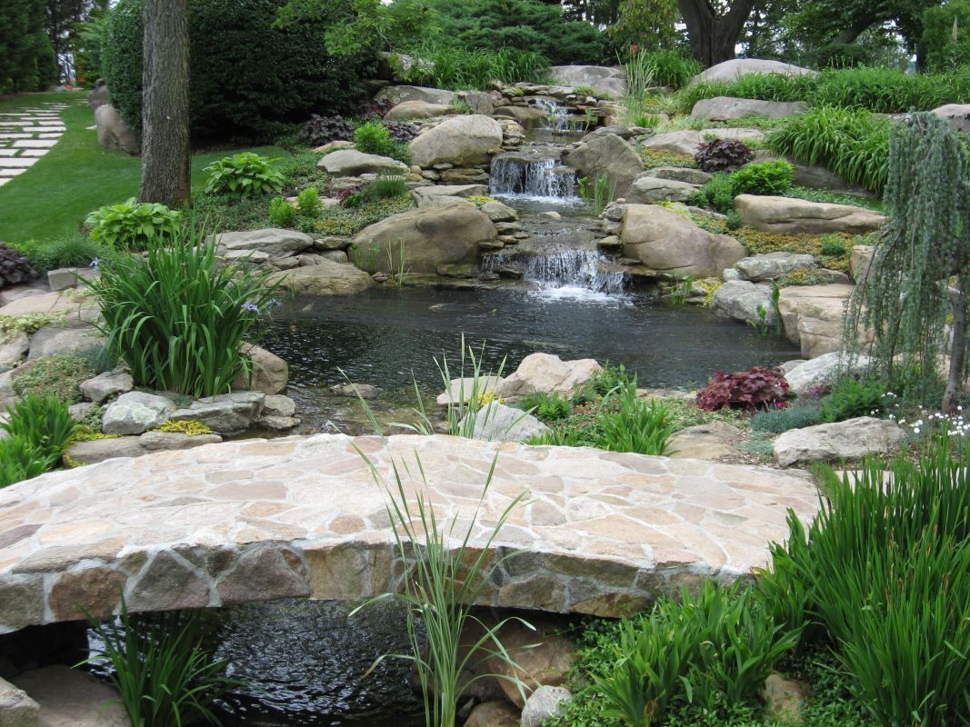 Stone bridge over a pond with a waterfall, lush greenery, and rocks