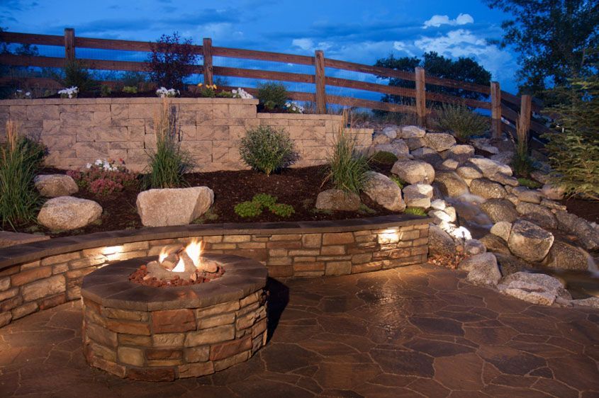 Stone fire pit with flames, surrounded by a stone patio and landscaping, at dusk