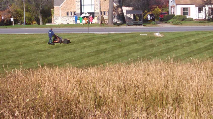 Person mowing grass near a road; a church and houses are in the background, golden field in the foreground