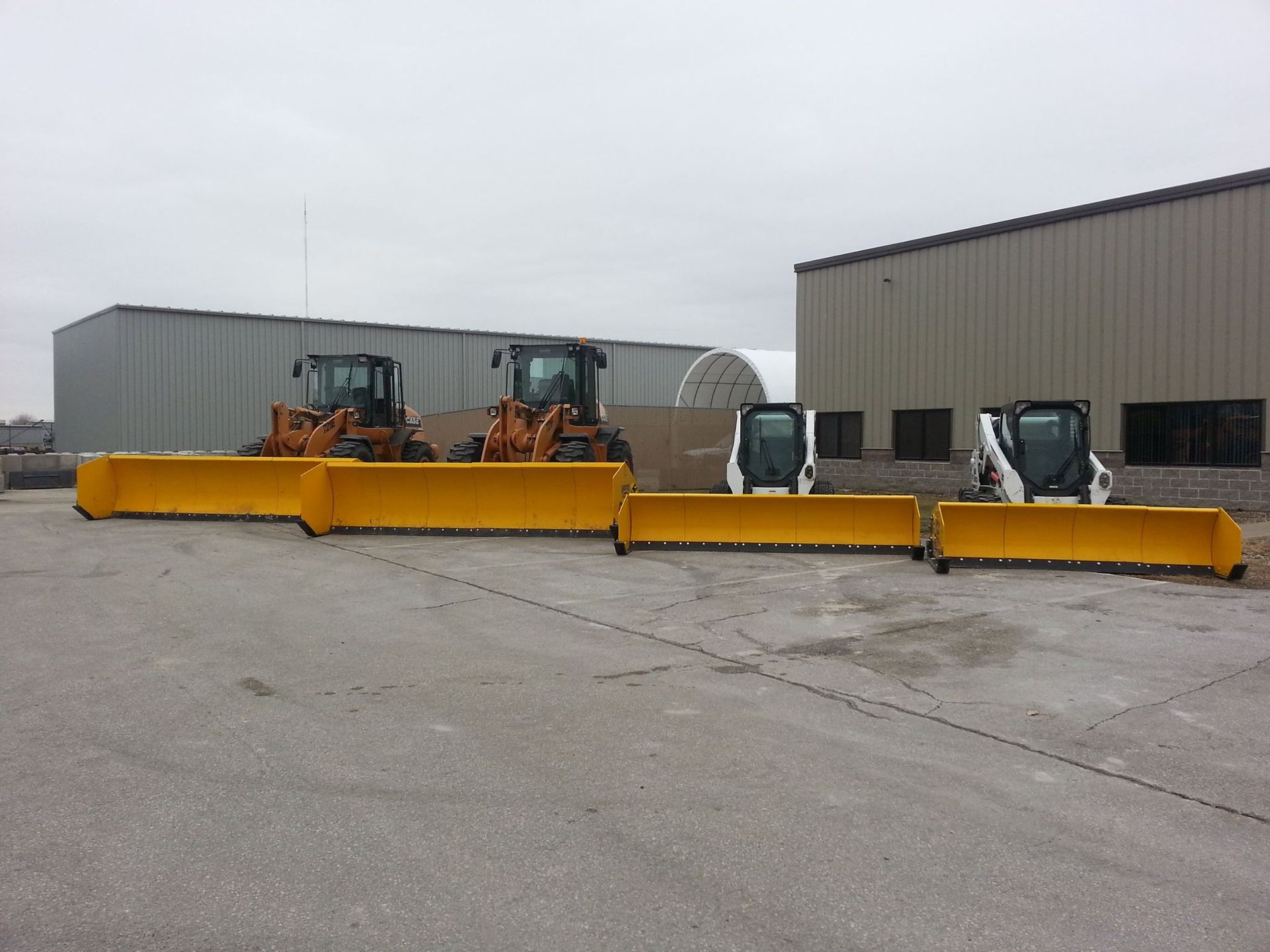 Three tractors with snowplows parked outside industrial buildings on a cloudy day