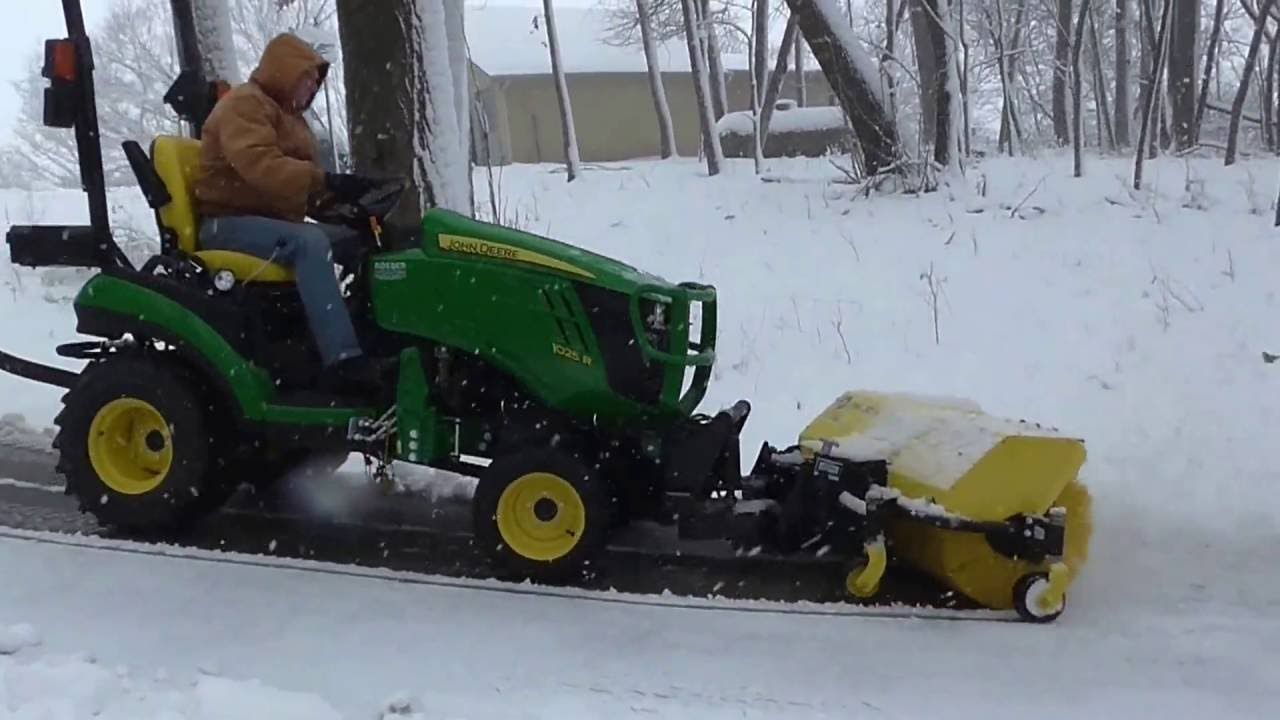 A person in a parka operates a green John Deere tractor with a snowblower on a snowy road
