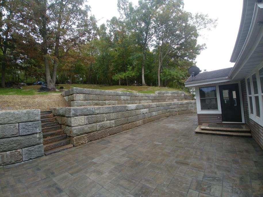 Stone retaining walls and steps lead to a patio next to a house with a black door.