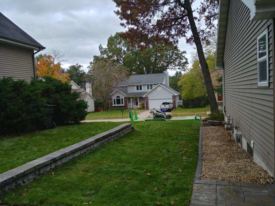 Green lawn and stone retaining walls in front yards, with houses and trees in the background under a cloudy sky.