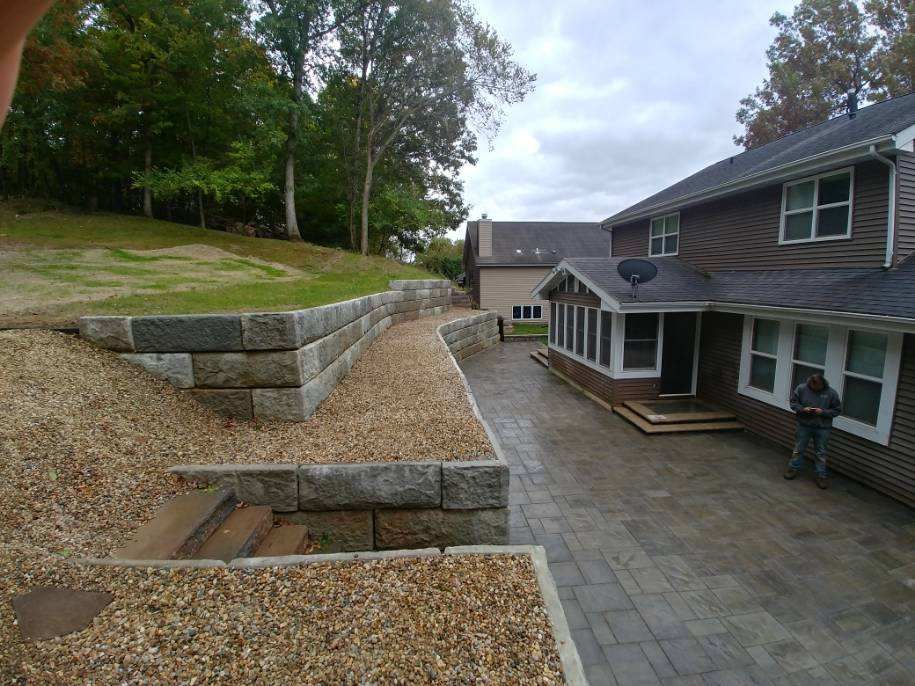 Stone retaining walls with gravel alongside a house with a patio and lawn on a cloudy day.