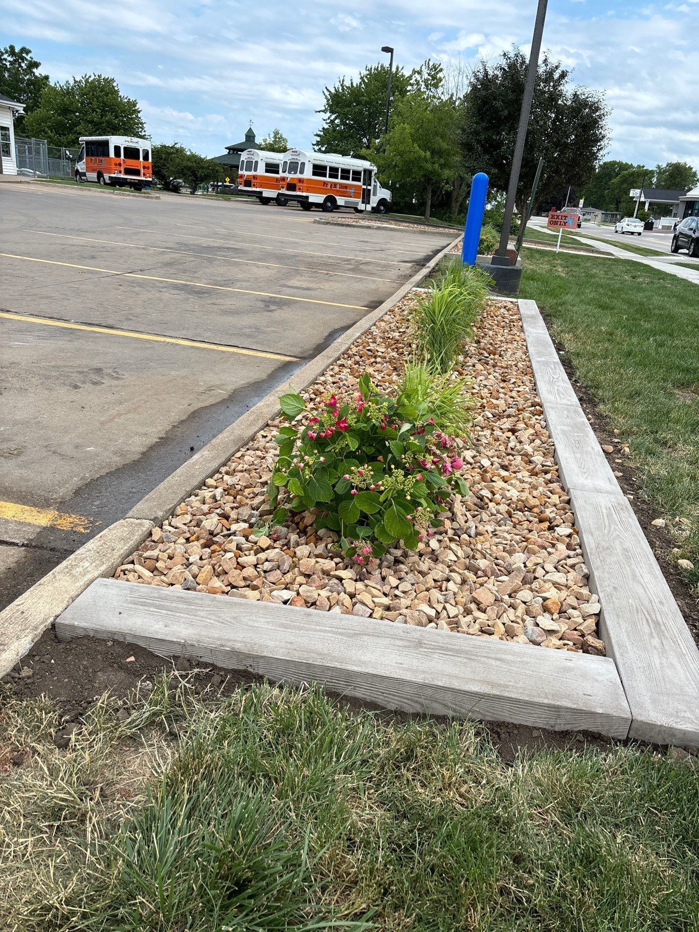 Stone-lined garden bed with plants and gravel, bordered by concrete on the edge of a parking lot.