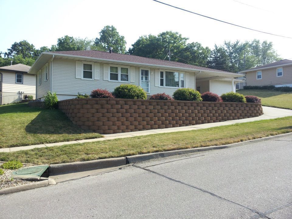 Beige ranch home with brown retaining wall and green shrubs along a sloping front yard next to a street.