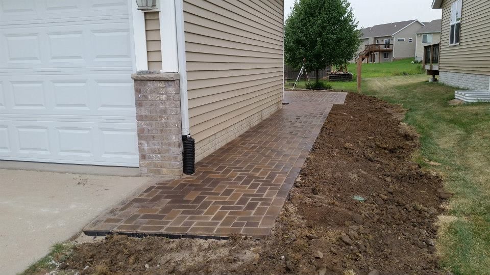 Brick pathway leading to a garage. Brown bricks, tan siding, and bare dirt.