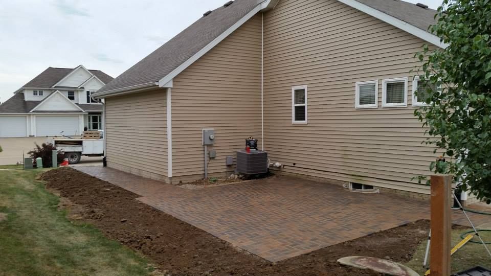 Newly constructed brick patio next to a tan-sided house. Brown earth borders the patio on a grassy lawn.