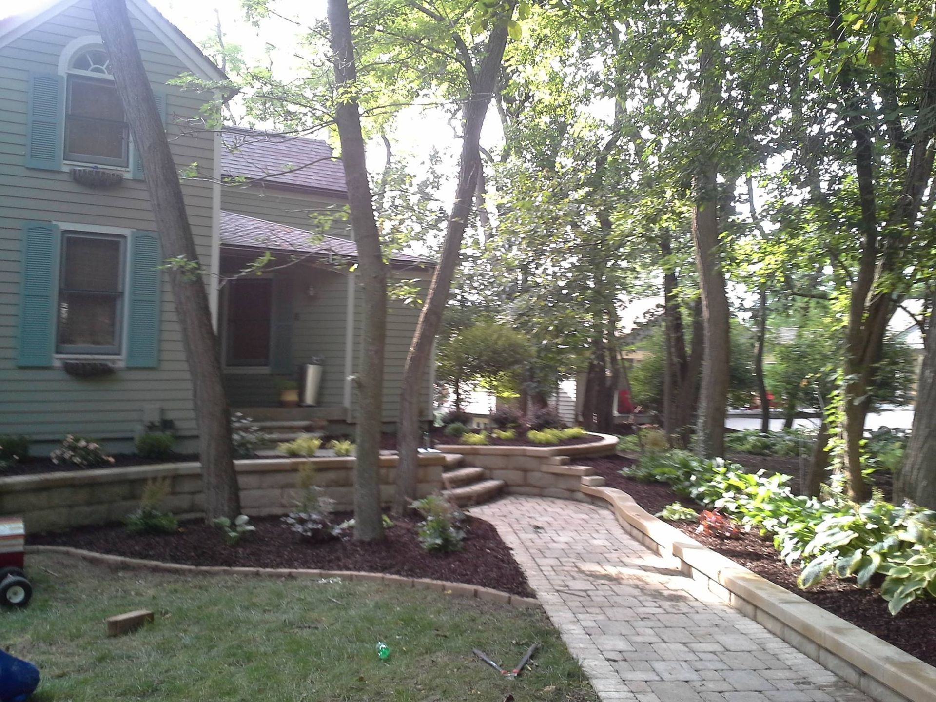 House with green shutters and trees, landscaped walkway with tiered retaining walls and flowers.