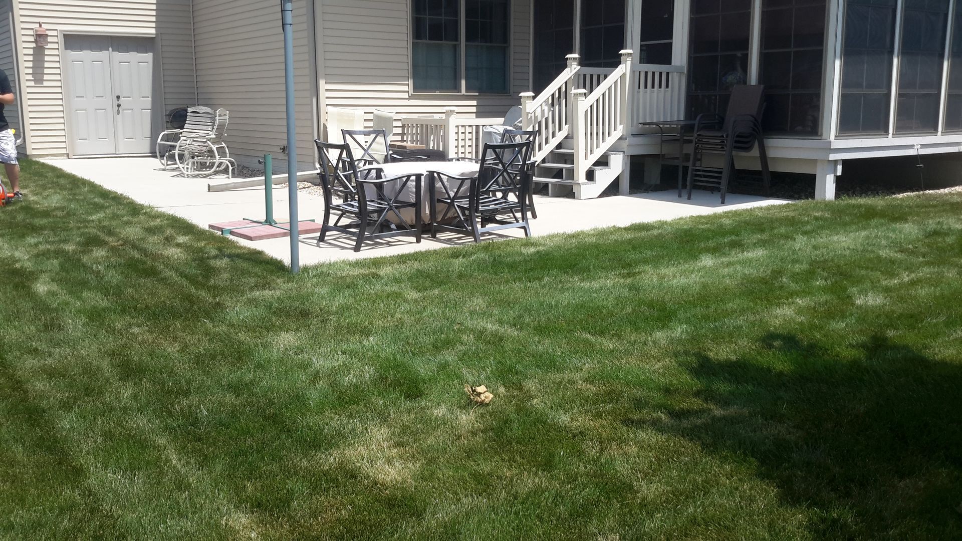 Lush green lawn slopes down to a concrete patio with a table and chairs. House in the background.