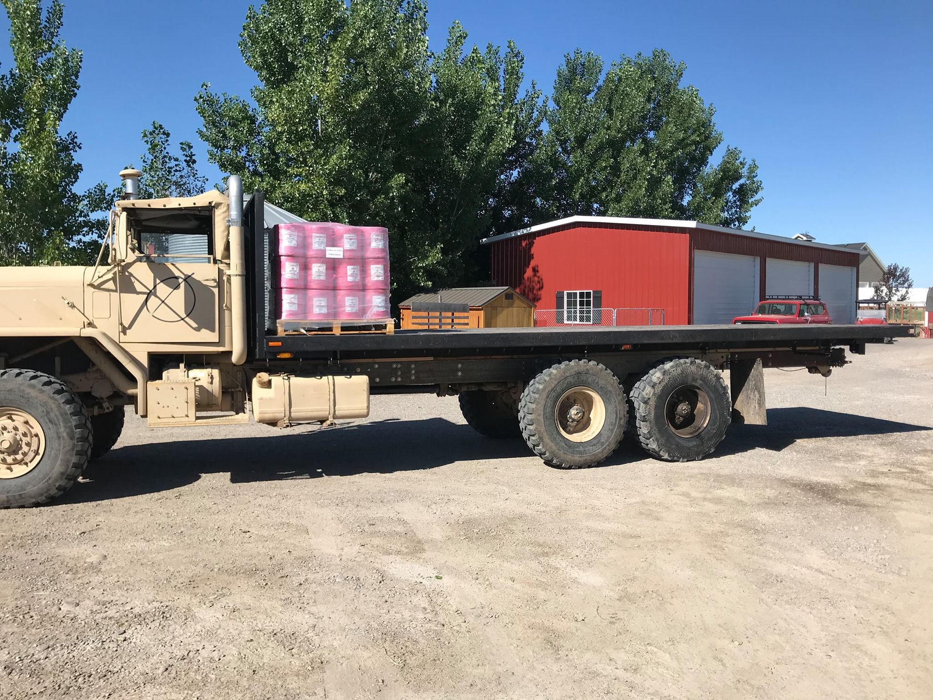 A military truck with a flatbed is parked in front of a red building.