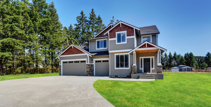 Two-story suburban house with garage, front porch, and large driveway surrounded by trees