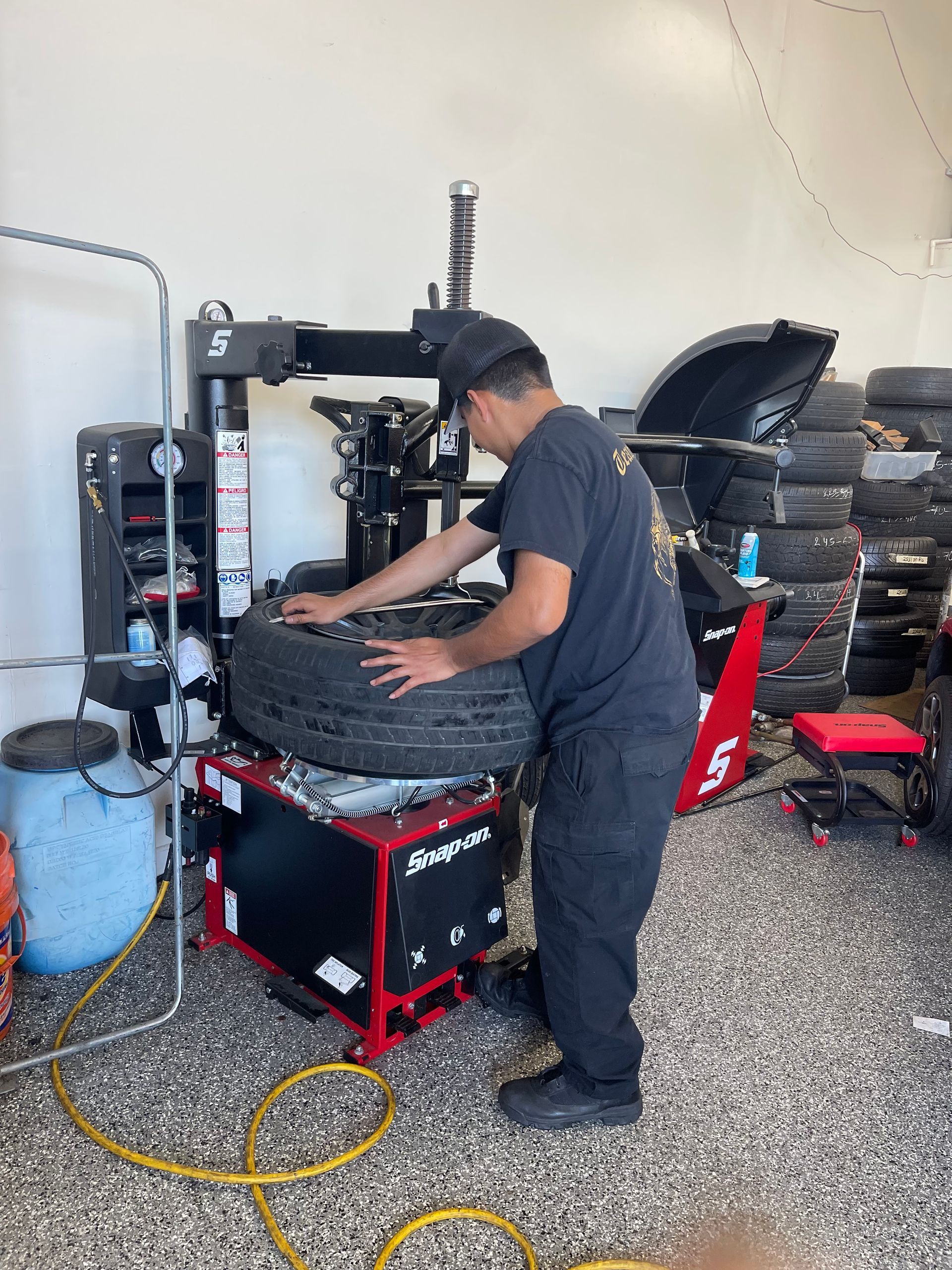 Mechanic changing a car tire on a tire machine in a garage with stacked wheels nearby.