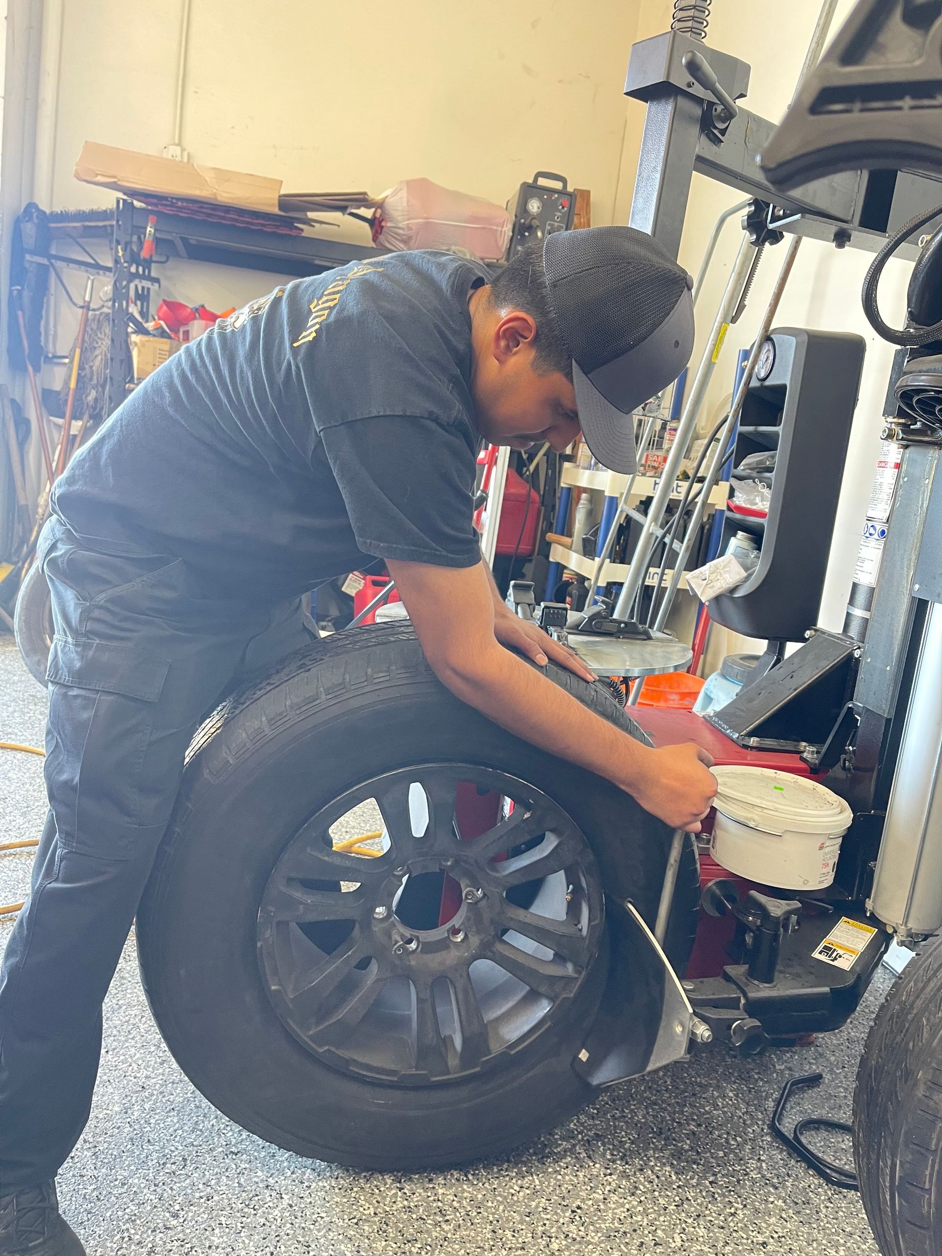 Mechanic crouches beside a car wheel in a garage, using tools to work on the brake assembly.