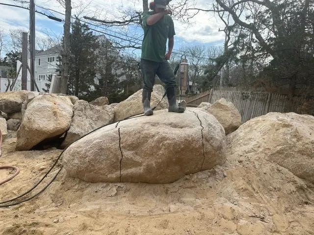 Man standing on a large boulder, using a tool to work on it. He is wearing a green shirt, and the setting is outdoors with other rocks visible.