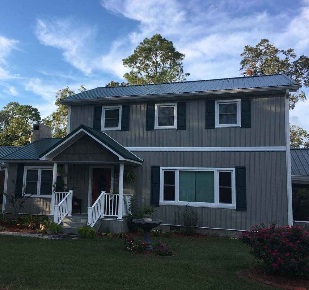 Two-story house with gray siding, green shutters, and a matching metal roof, set against a blue sky.