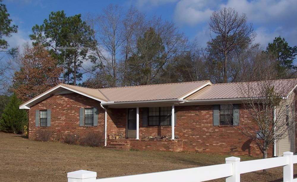 Red brick house with metal roof, small porch, and white fence in front. Trees in background.