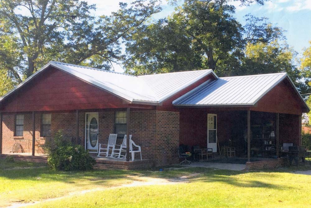 Red brick and wood house with a metal roof and porch, on a grassy lawn.