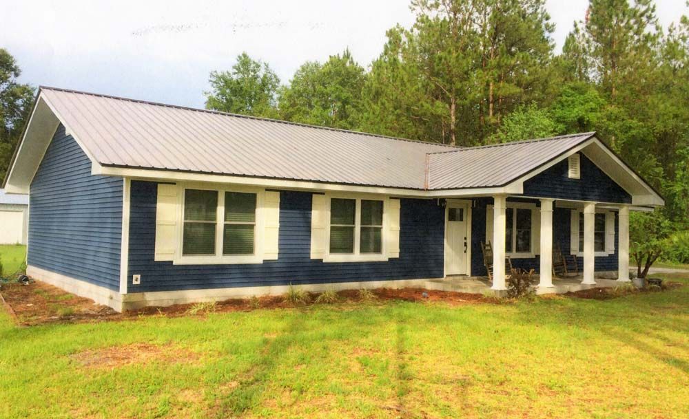 Blue house with metal roof, white trim, and a porch with columns, set on a grassy lot.