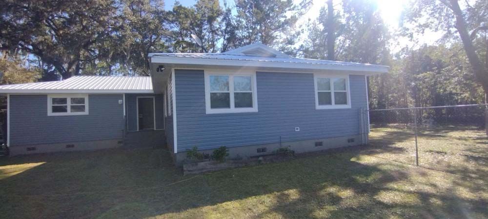 Blue-sided house with white trim, windows, and roof on a grassy yard. Trees in the background.