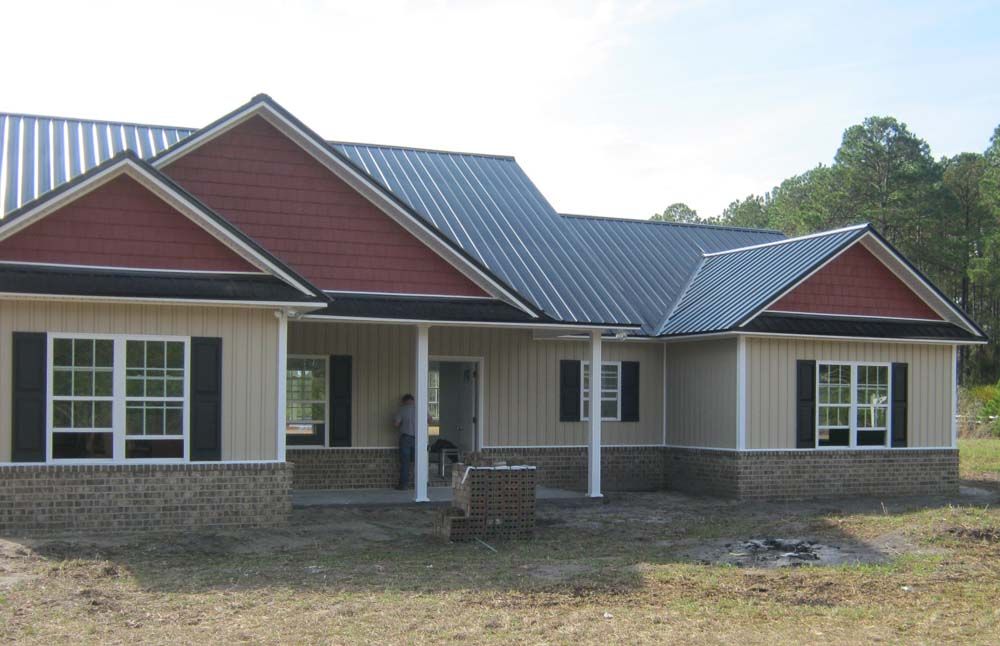 Tan house with a red roof, black shutters, and a metal roof on a sunny day.