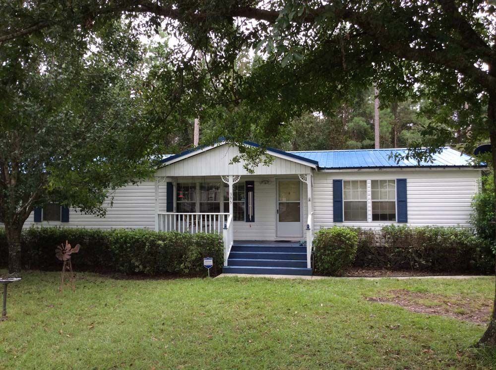 White mobile home with blue roof, door, and shutters; porch with white railing; front yard with bushes and trees.