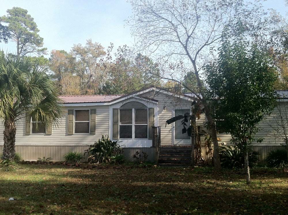 A weathered, one-story house with tan siding, a red roof, and a small porch, surrounded by trees.