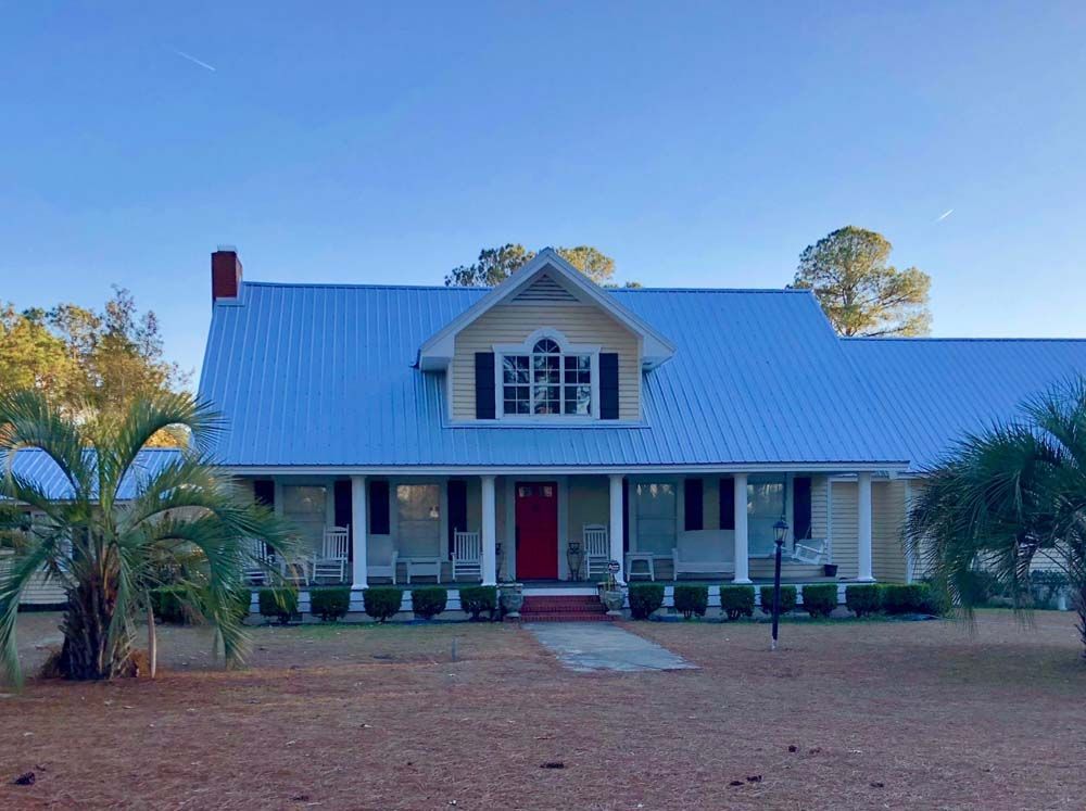 Yellow and white farmhouse with blue metal roof, red door, and porch with white rocking chairs.