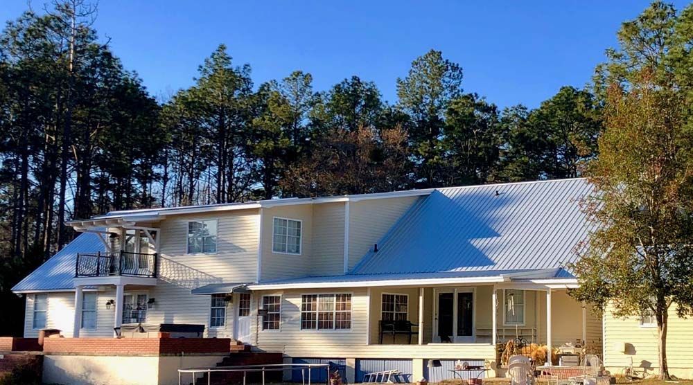Large, light yellow house with a white metal roof, porch, and balcony against a backdrop of tall green trees and a blue sky.