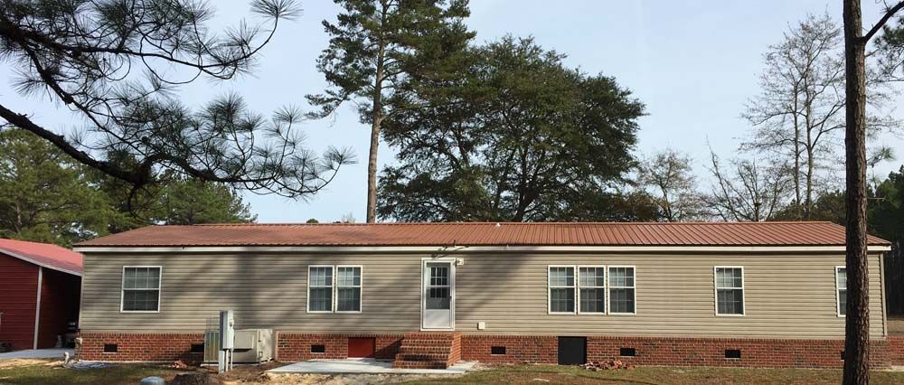 Tan mobile home with a red metal roof, brick foundation, and trees in the background.