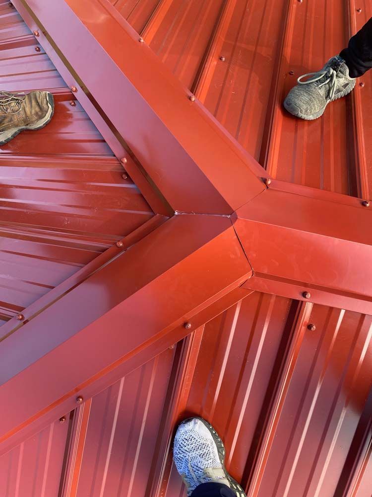 Red metal roof with intersecting ridges; two people's feet visible.