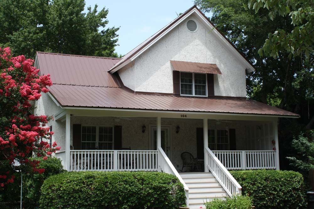 White house with brown metal roof, porch, and landscaping.