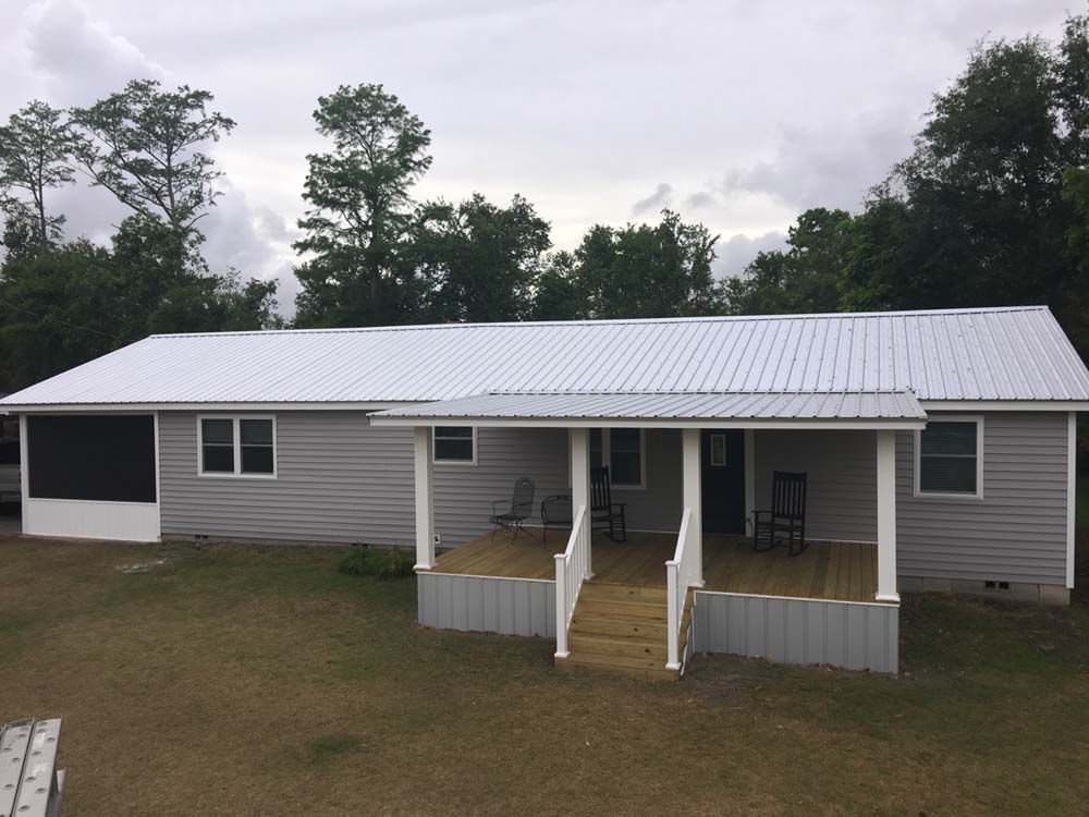 Gray-sided house with a white metal roof and covered porch in a grassy yard under a cloudy sky.