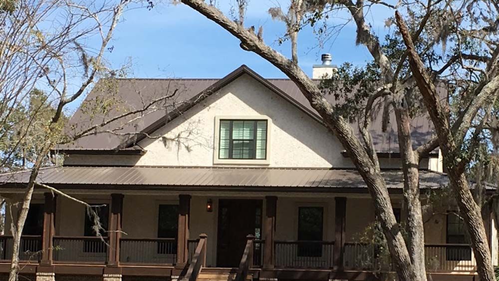 Tan house with brown metal roof, porch, and trees.