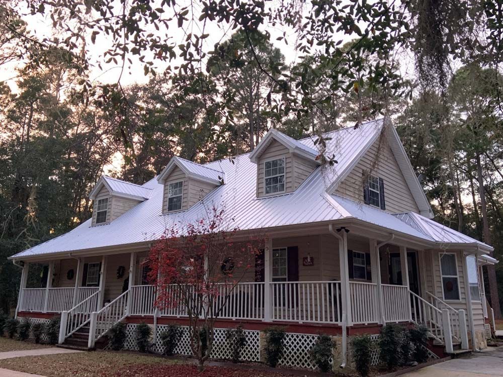 A two-story house with a white wraparound porch, metal roof, and dormer windows, surrounded by trees.